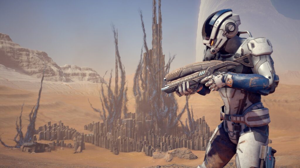 Ryder, de armadura branca e azul e capacete, aponta um rifle num deserto com estruturas alienígenas ao fundo, em cena de Mass Effect: Andromeda