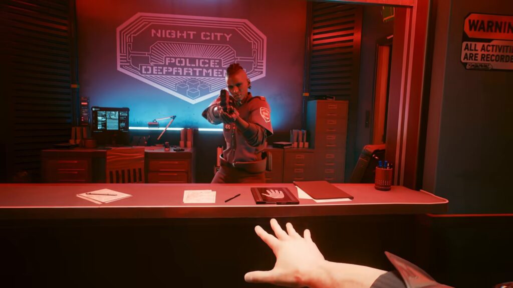 Uma policial aponta a arma para V atrás do balcão de uma delegacia da Polícia de Night City (NCPD), iluminada de vermelho, em cena do vídeo oficial da atualização 2.5 de Cyberpunk 2077