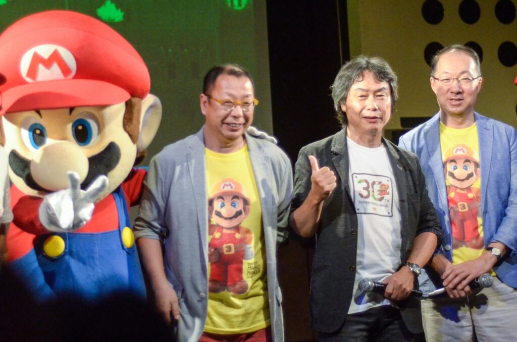 Takashi Tezuka, Shigeru Miyamoto, com o polegar para cima, e Koji Kondo no palco da festa de 30 anos de Super Mario Bros., em Tóquio, ao lado de um Mario de pelúcia gigante