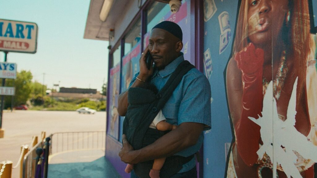 Mahershala Ali carrega um bebê num canguru enquanto fala ao telefone na calçada
