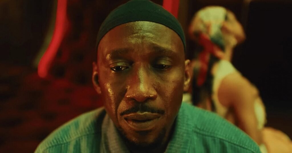 Mahershala Ali, de gorro verde, em close sob luz vermelha em Your Mother Your Mother Your Mother