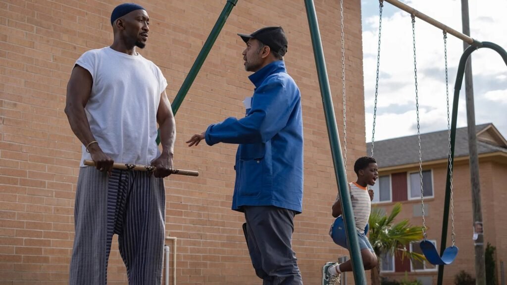 Mahershala Ali, segurando um bastão, conversa com um homem num parquinho, com um menino no balanço ao fundo